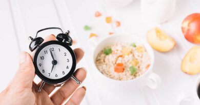 A person's hand holding alarm clock in hand over the breakfast on the table. intermittent fasting