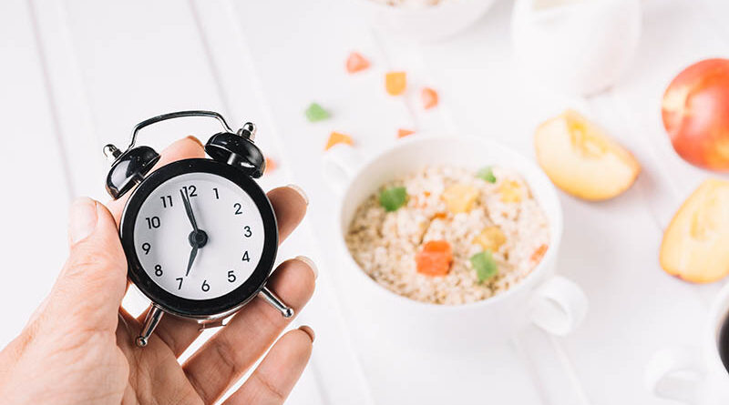 A person's hand holding alarm clock in hand over the breakfast on the table. intermittent fasting