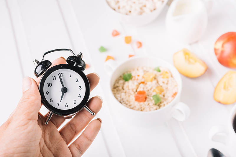 A person's hand holding alarm clock in hand over the breakfast on the table. intermittent fasting